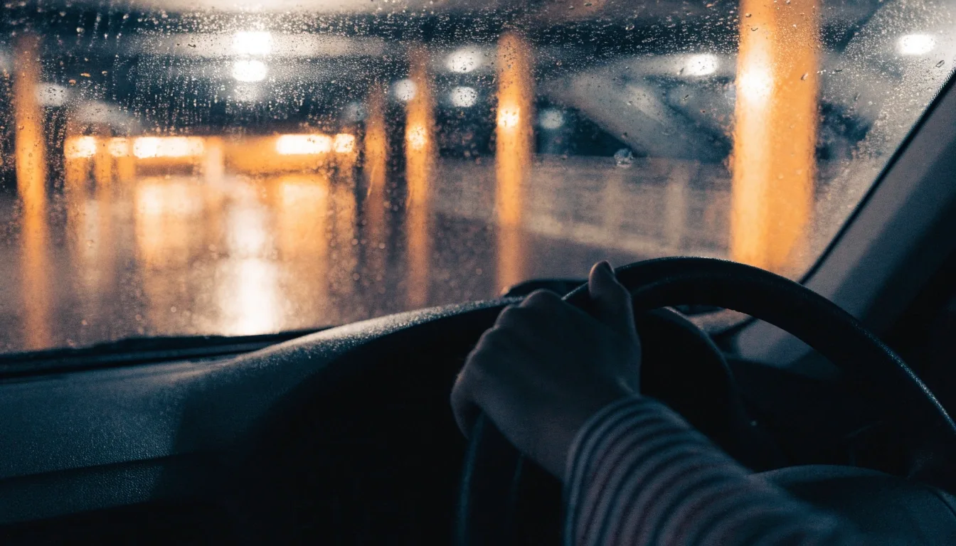Rain-beaded windscreen at night with warm car park bay lighting behind the glass