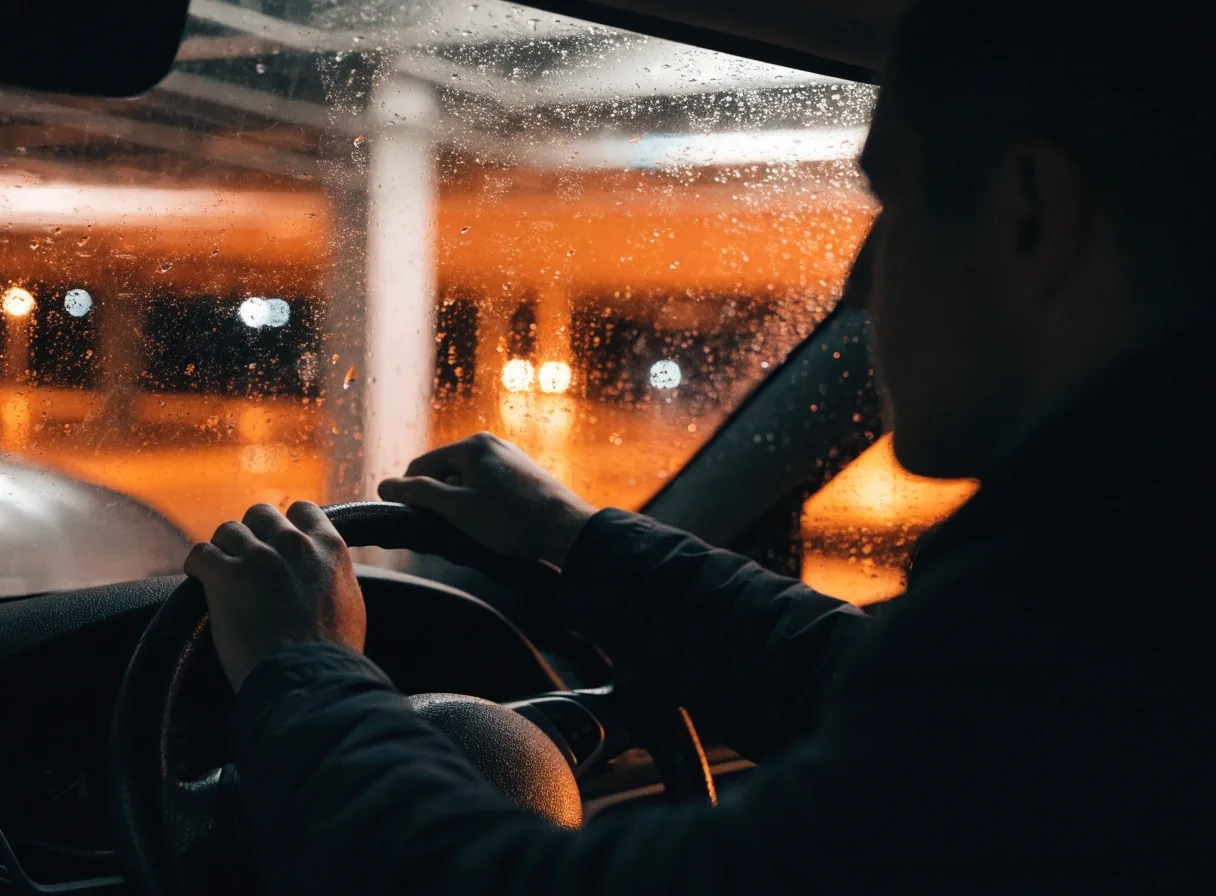 Hands on a steering wheel inside a covered car park with rain on the windscreen at night